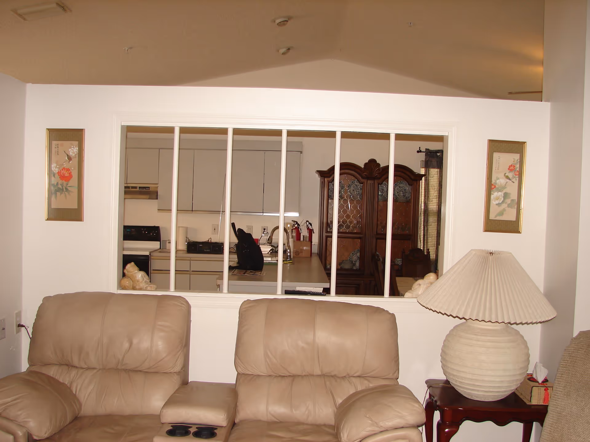 Interior view of a living room with two beige leather recliners in the foreground. Behind a half wall with vertical bars, a kitchen and dining area are visible, including white cabinets, a stove, a wooden china cabinet, and a dining table. A large beige lamp sits on a wooden side table next to the recliners. Two framed floral artworks hang on the wall on either side of the half wall.