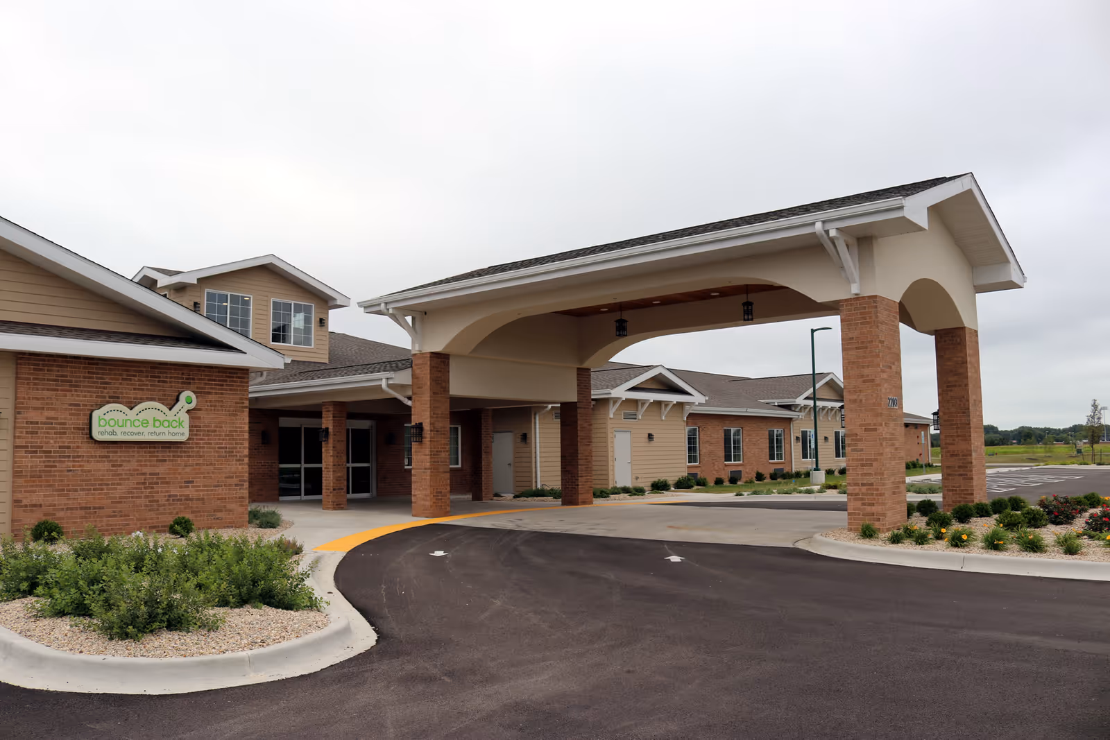 Covered porte-cochère and main entrance of a brick-faced senior living building with landscaping and signage.