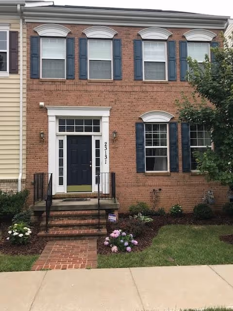 Front exterior view of a two-story brick townhouse with a black front door, white trim, and blue shutters on the windows. There are steps leading up to the entrance with black railings, a small garden with flowers and shrubs, and a sidewalk in front.