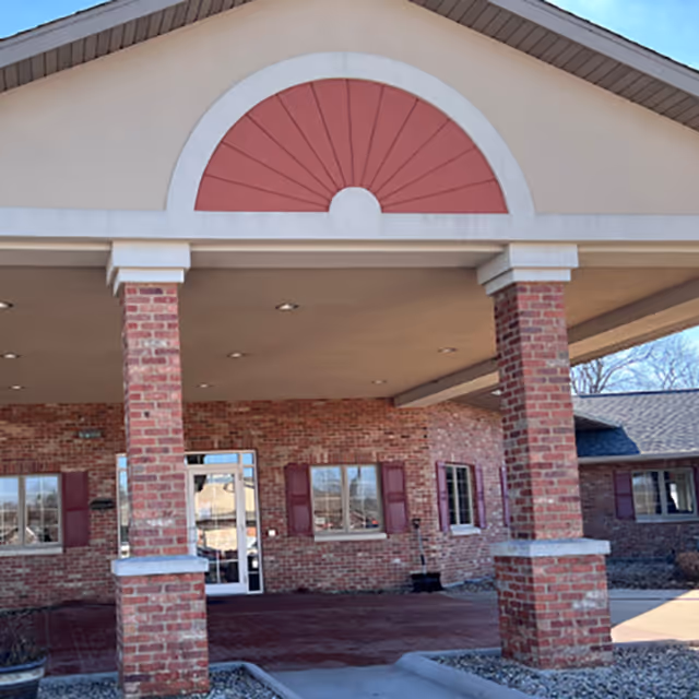 Front exterior view of a brick building with a covered entrance supported by two brick columns. The building has windows with red shutters and a decorative arch above the entrance.