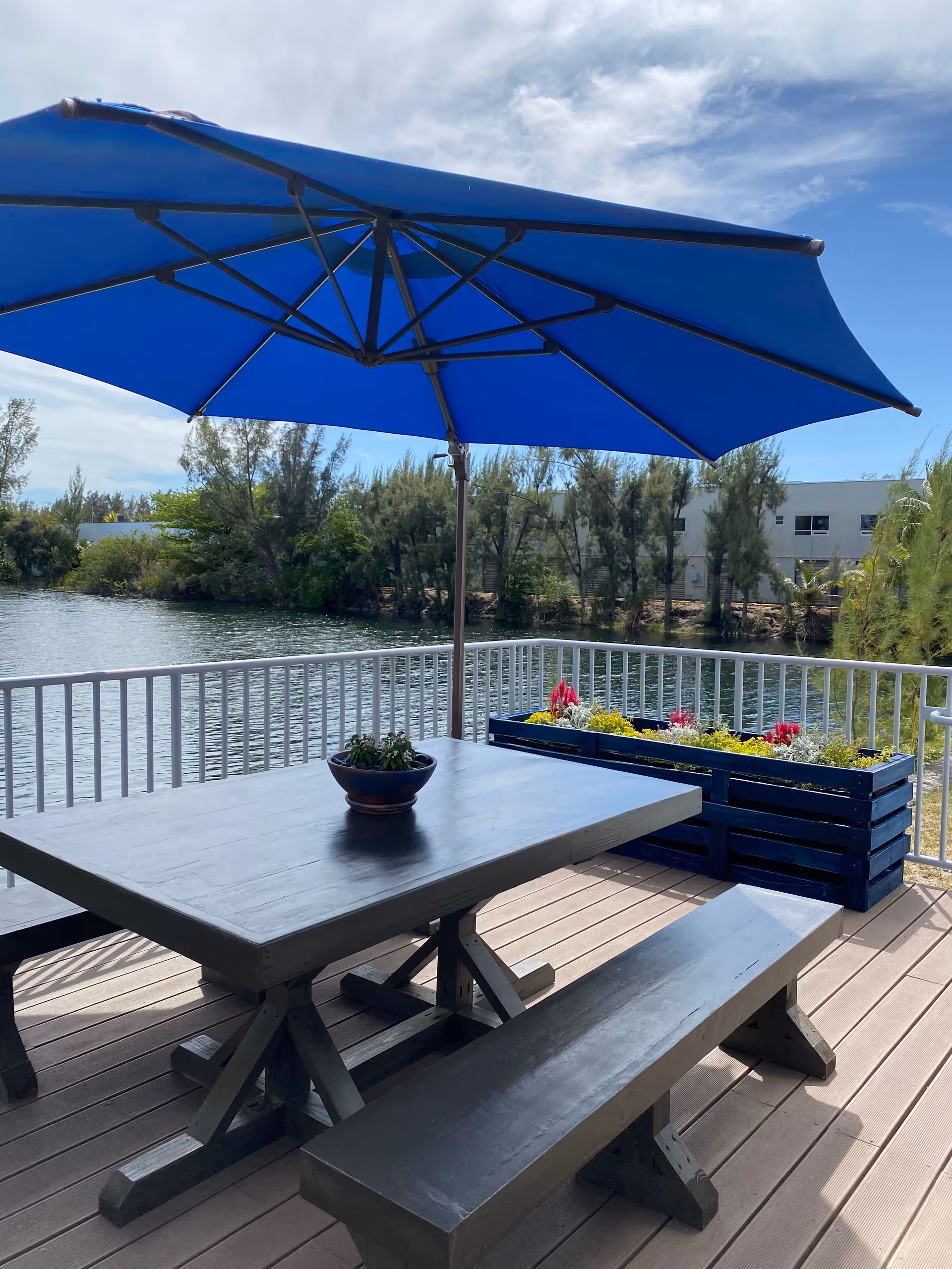 Outdoor wooden picnic table under a large blue umbrella on a waterfront deck.