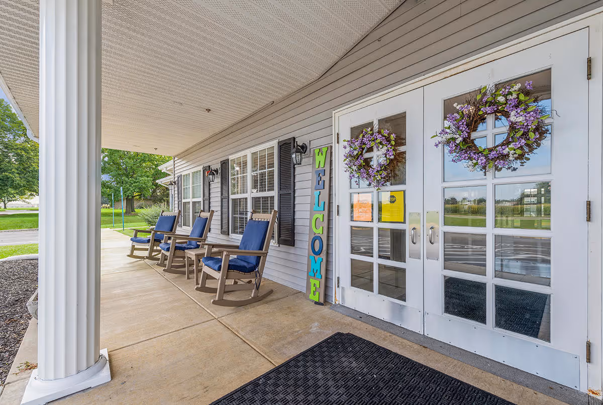 Front porch of Digby Place featuring a covered walkway with white columns, three wooden rocking chairs with blue cushions, a vertical colorful 'WELCOME' sign, and double glass doors adorned with purple floral wreaths. Green trees and a grassy area are visible in the background.