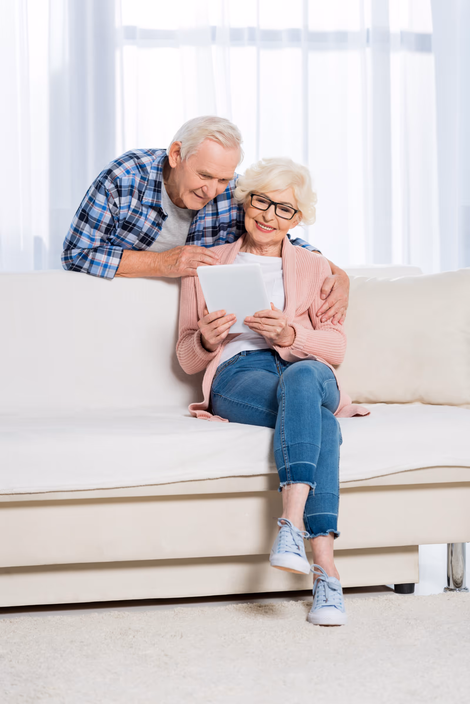 Senior couple sits on a bright living room sofa, looking at a tablet.