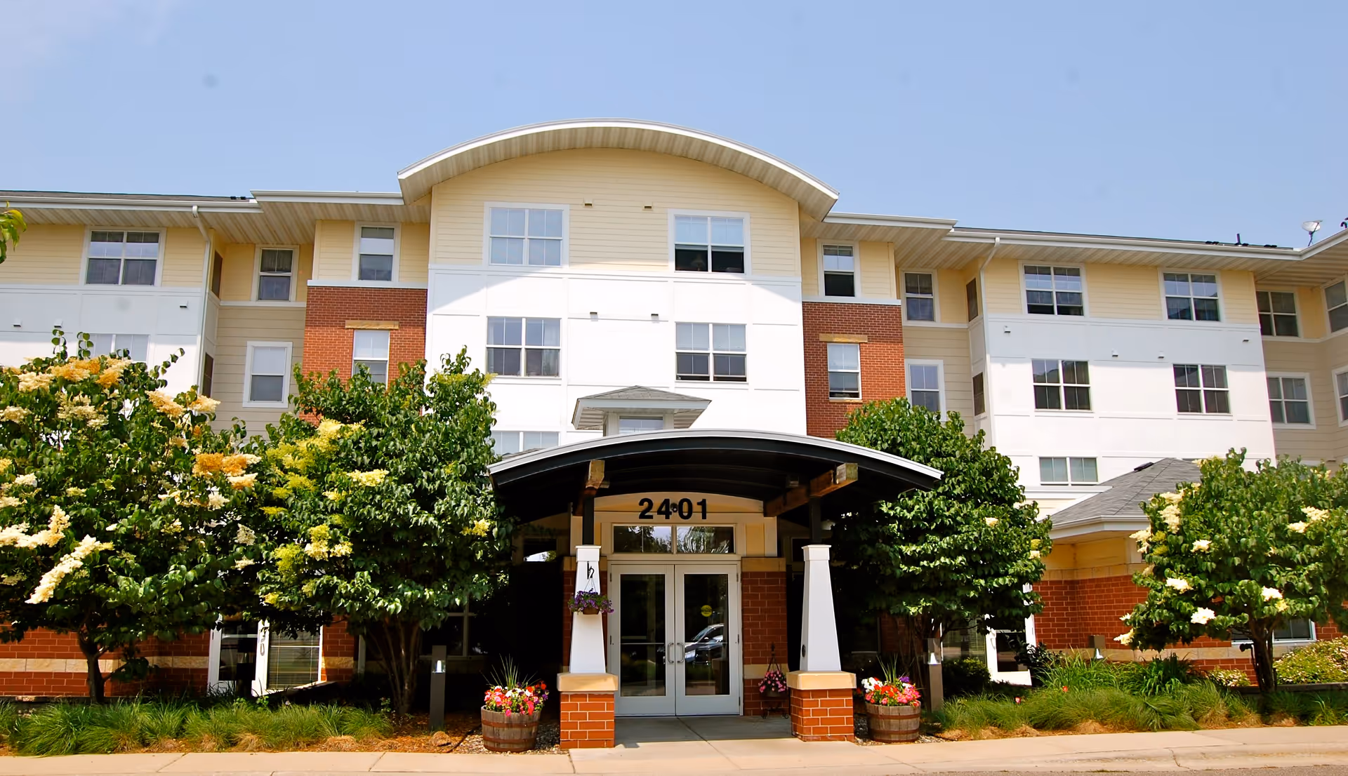 Front entrance of a four-story senior apartment building with a covered portico, address number 2401, and flowering trees.