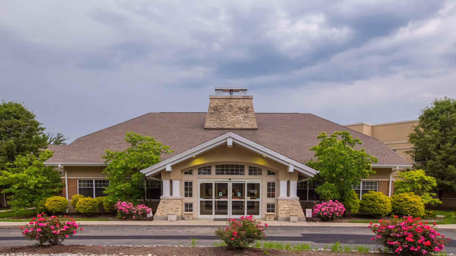 Front exterior view of a single-story building with a peaked roof and stone chimney feature. The entrance has double glass doors under a covered porch supported by white columns. The building is surrounded by green trees, bushes, and blooming pink flowers, with a cloudy sky overhead.