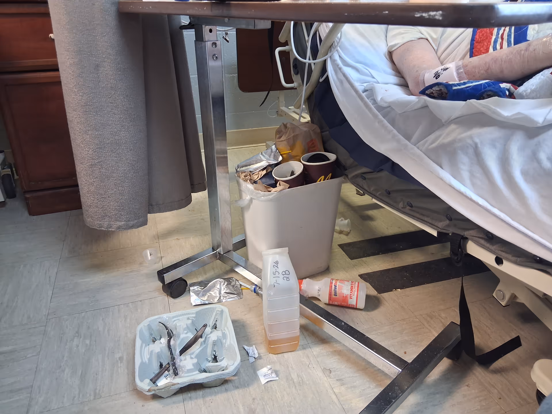 Hospital bed in a patient room with cups and scattered supplies nearby.