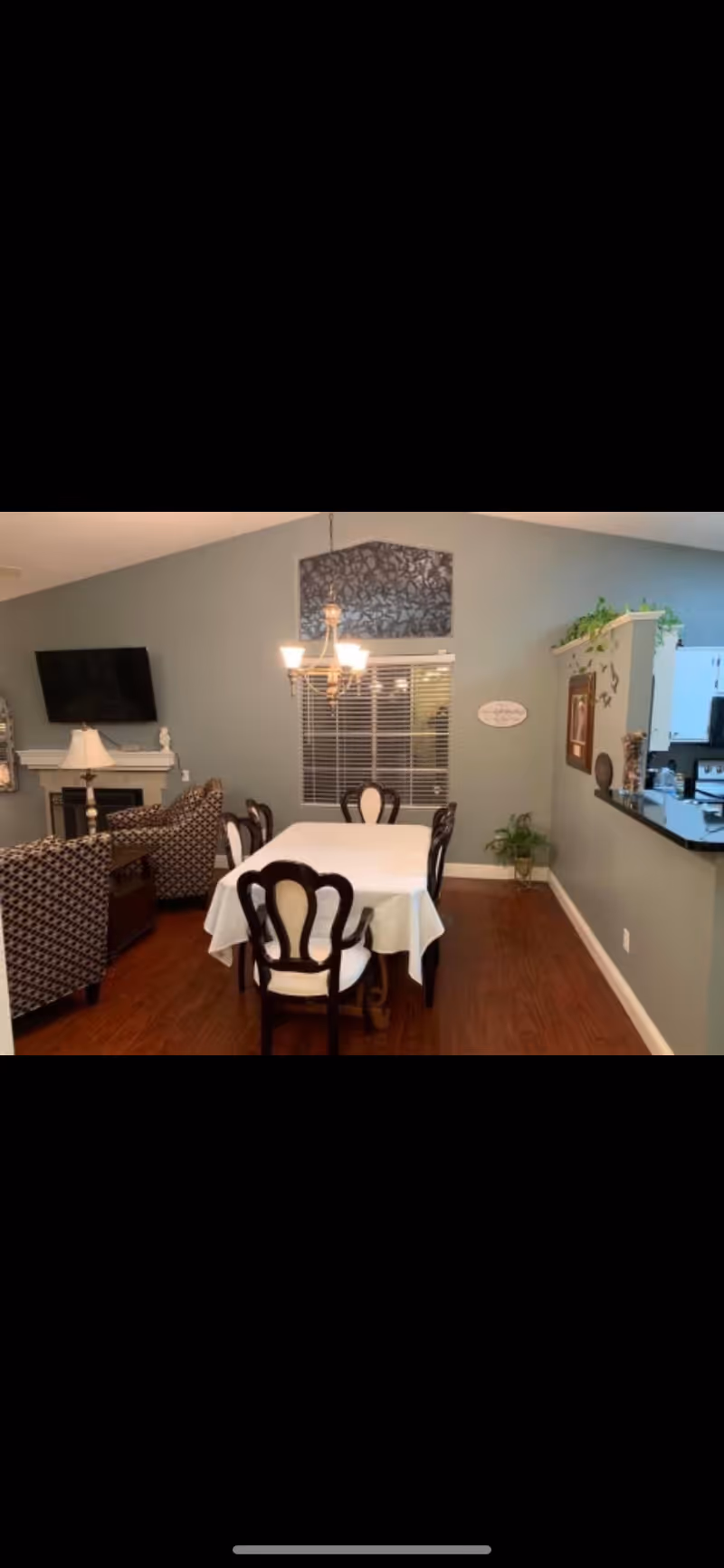A dining room with a rectangular table covered with a white tablecloth and six dark wooden chairs with white cushions. A chandelier with five lights hangs above the table. In the background, there is a window with blinds and a decorative wall hanging above it. To the left, there is a living area with patterned armchairs, a fireplace, and a wall-mounted TV. To the right, there is a partial view of a kitchen with a counter and some plants on top.