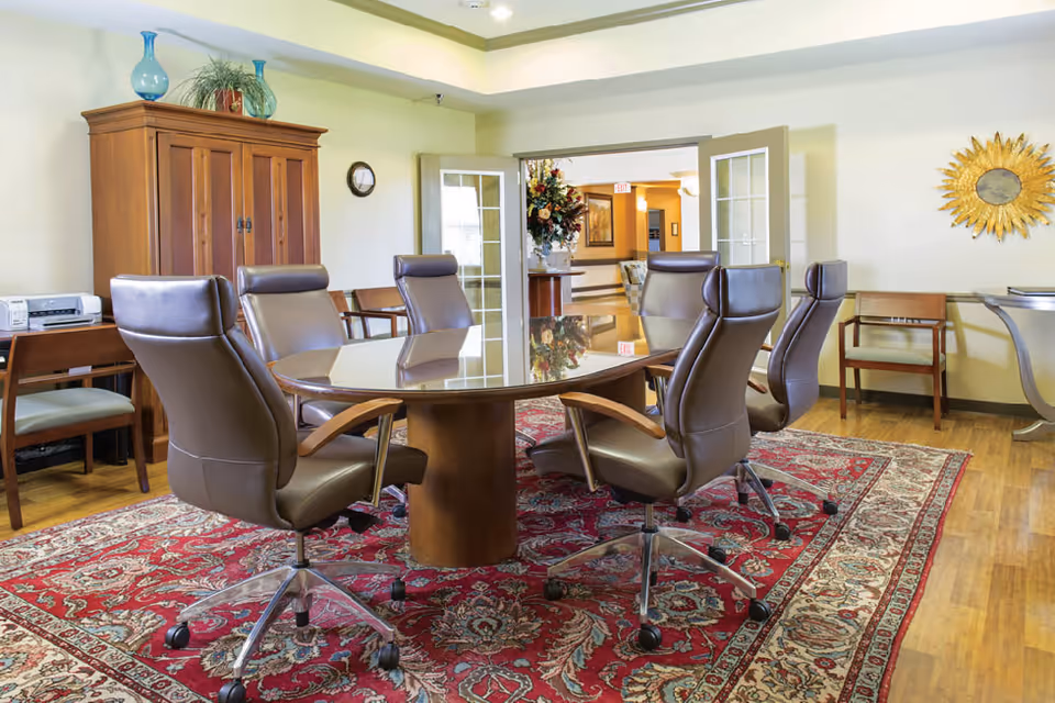 A conference or meeting room with a large oval wooden table surrounded by six brown leather swivel chairs on a red patterned rug. The room has wooden flooring, a wooden cabinet with decorative vases on top, a small desk with a printer, and a wall clock. French doors open to a hallway decorated with flowers and artwork. A sunburst mirror hangs on the wall.