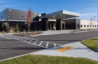 Exterior view of a modern senior living facility building with a covered entrance, parking area, and landscaped surroundings under a clear sky.