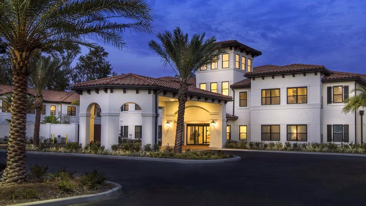 Exterior view of a senior living facility building at dusk with palm trees in front, warm lights glowing from windows, and a covered entrance with arches.