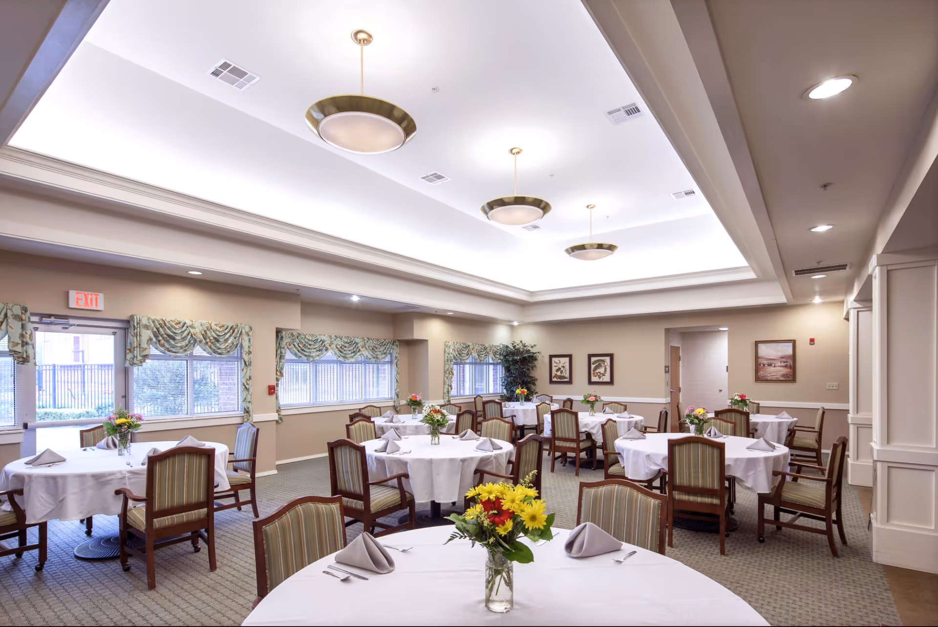 A bright and spacious dining room with round tables covered in white tablecloths, each set with folded napkins, silverware, and a vase of colorful flowers. The room has large windows with patterned valances, beige walls, framed artwork, and ceiling lights with brass fixtures.