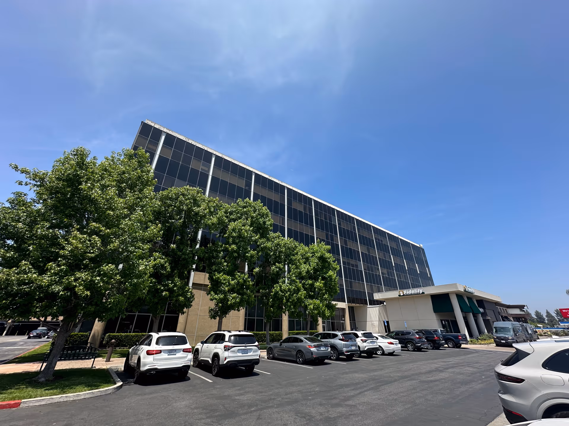 Glass-front building with parked cars and trees under a blue sky.