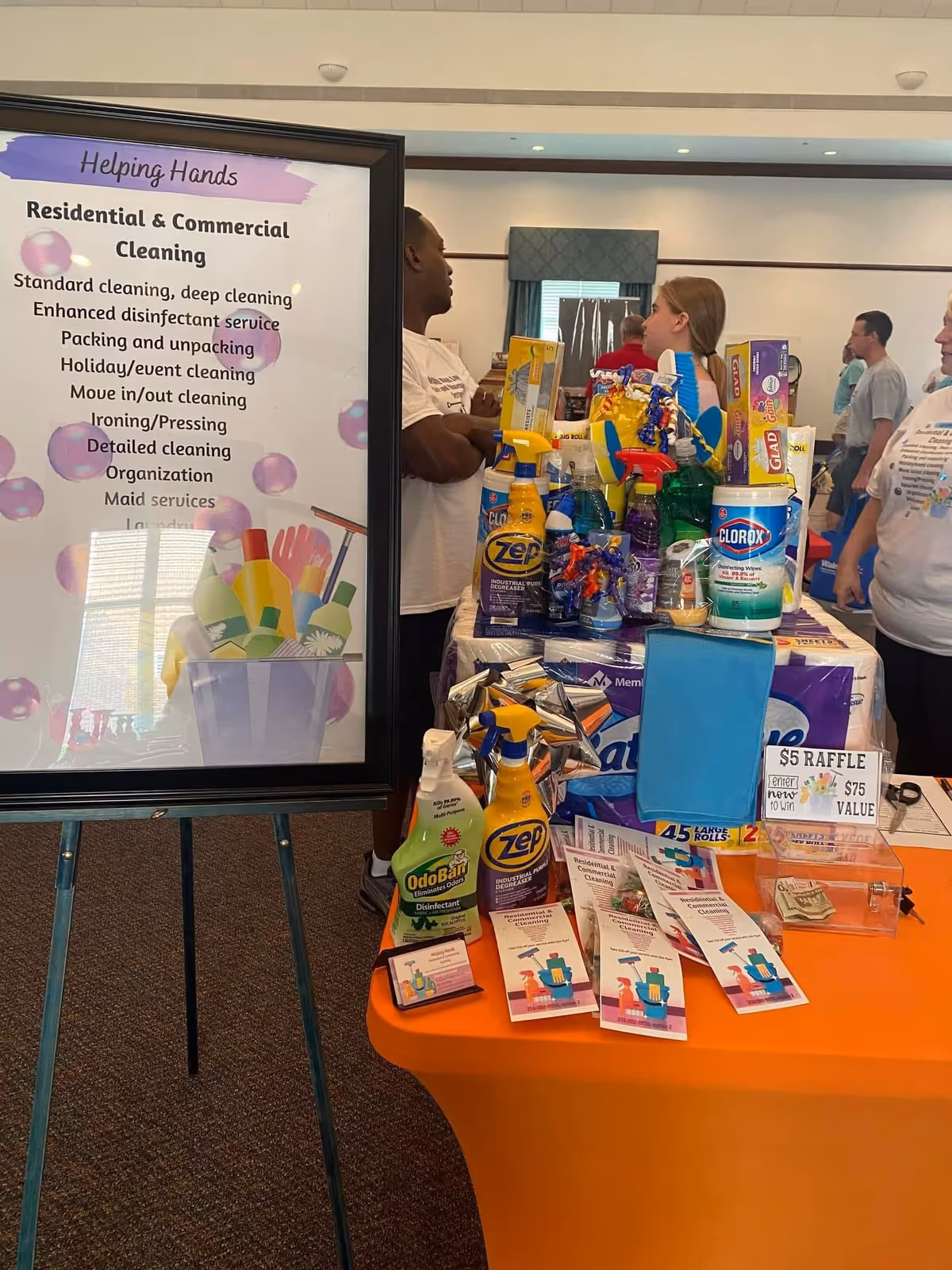 Indoor display table of cleaning supplies beside a Helping Hands sign at an event