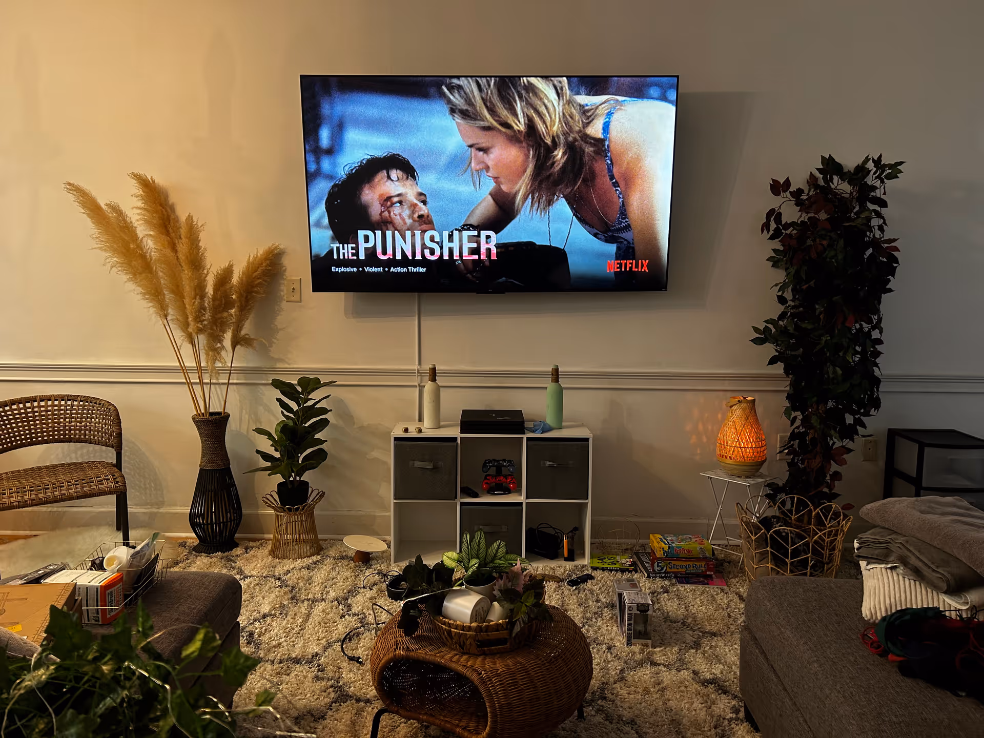Living room with wall-mounted TV showing The Punisher, wicker furniture, plants, and a rug.