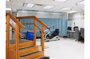 Interior exercise room in a senior facility with machines, a wooden railing, and curtains.