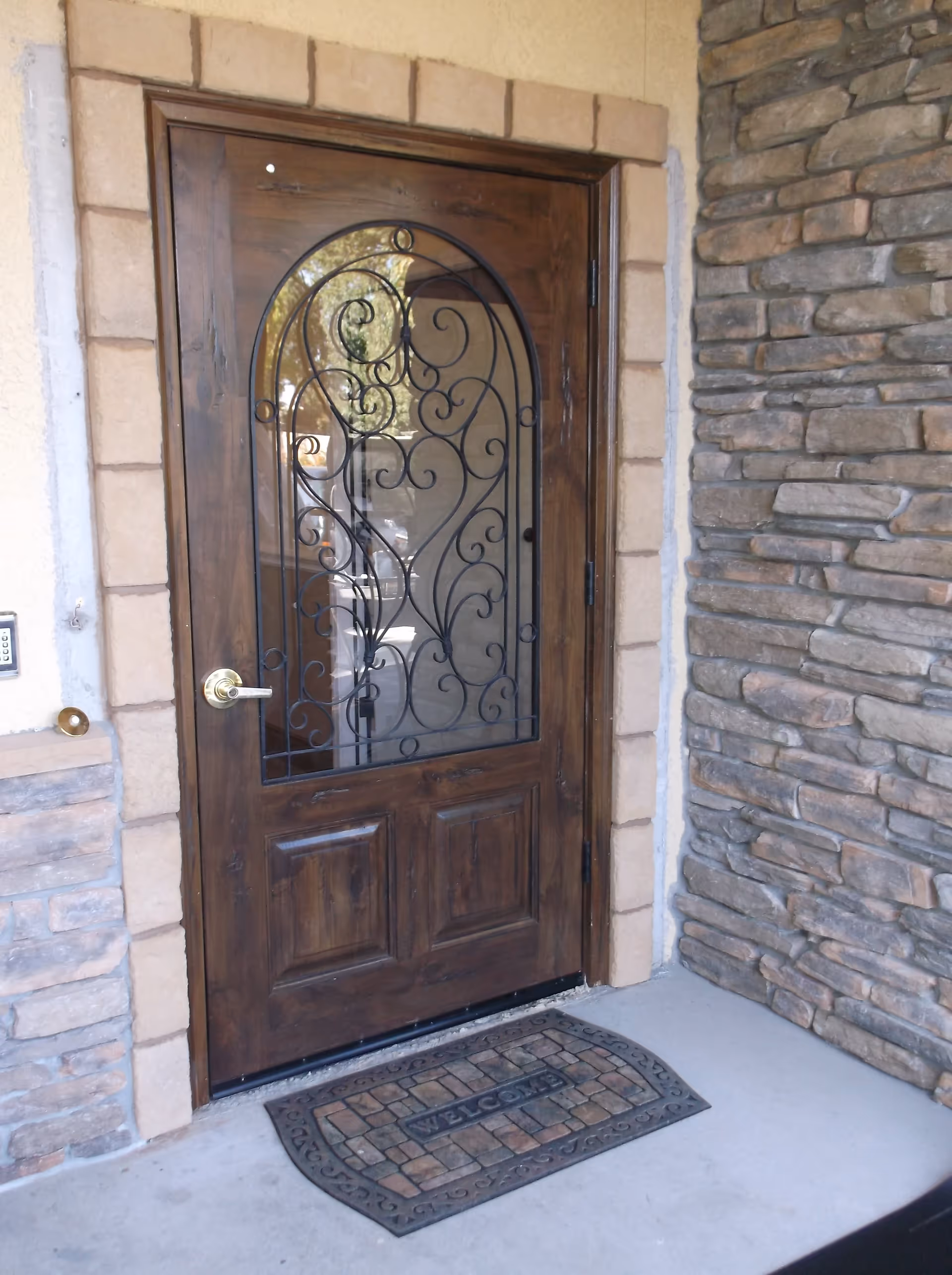 A wooden front door with decorative wrought iron design on the glass panel. The door is framed by stone and brick walls, and there is a welcome mat on the concrete floor in front of the door.