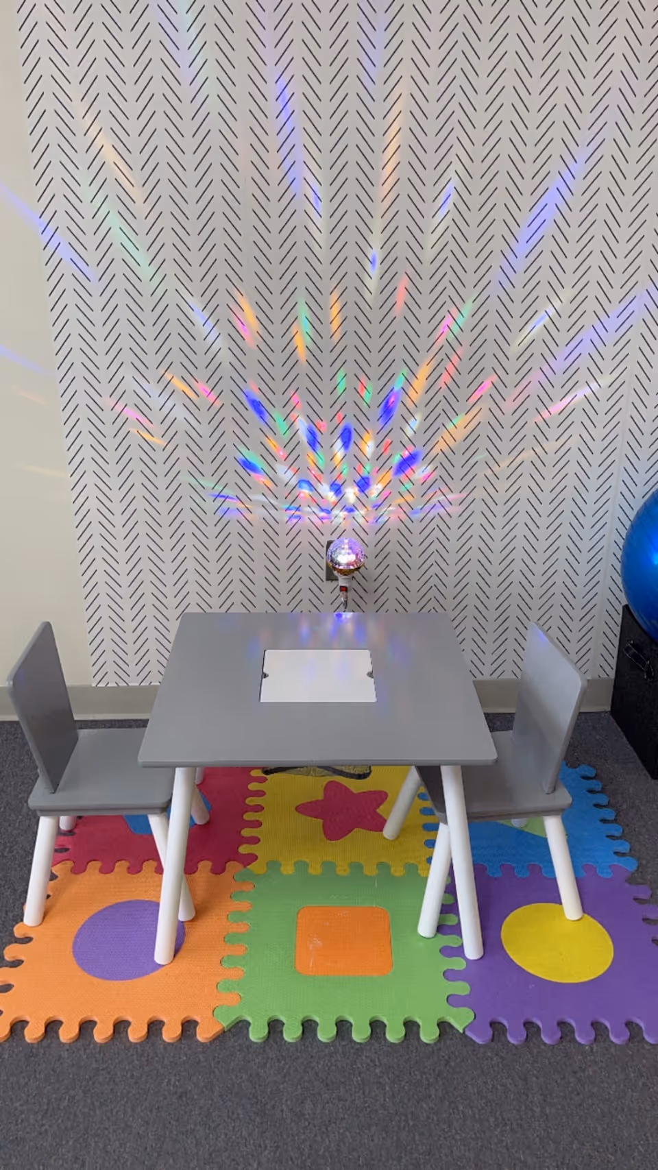 Small gray table with two chairs on colorful foam mats; rainbow light from a disco ball.