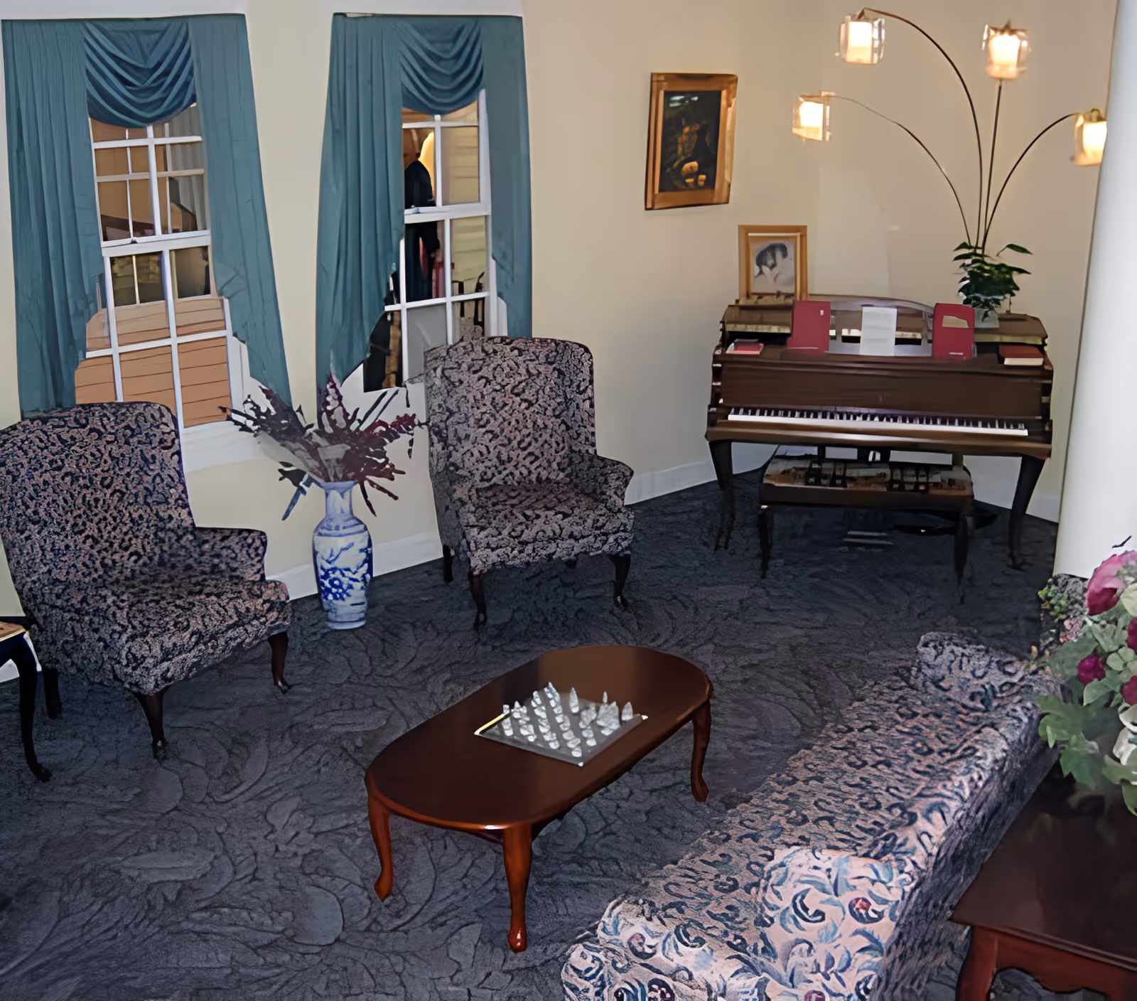 A cozy living room with patterned upholstered chairs and a matching sofa arranged around a wooden coffee table with a chess set on it. There is a piano against the wall with sheet music and framed pictures on top. The room has blue curtains on the windows and a tall floor lamp with multiple lights. A vase with flowers is placed near the window.
