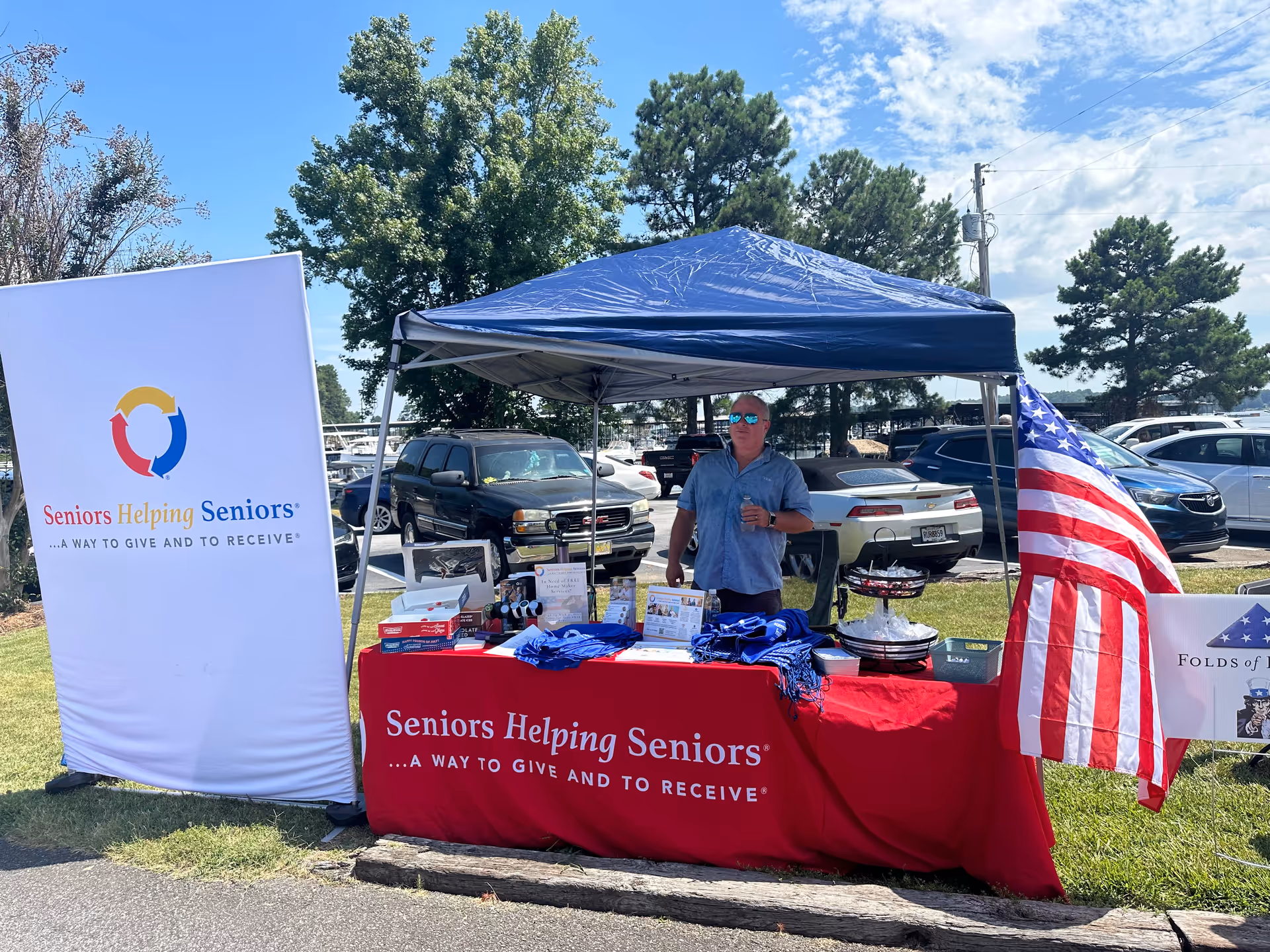 Outdoor booth for Seniors Helping Seniors in a parking lot with banners, table, and flag.