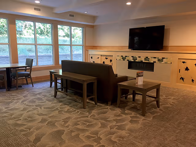 A cozy common area with a sofa and two wooden tables in front of a fireplace with decorative tiles. Above the fireplace is a mounted flat-screen TV. Large windows on the left side let in natural light, and there are additional chairs and tables near the windows. The room has a patterned carpet and soft lighting from ceiling fixtures.