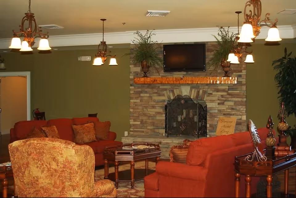 A cozy living room with red upholstered sofas and a patterned armchair arranged around a wooden coffee table. The room features a stone fireplace with a mounted flat-screen TV above it, flanked by two decorative plants. Warm lighting is provided by multiple hanging chandeliers, and the walls are painted olive green.