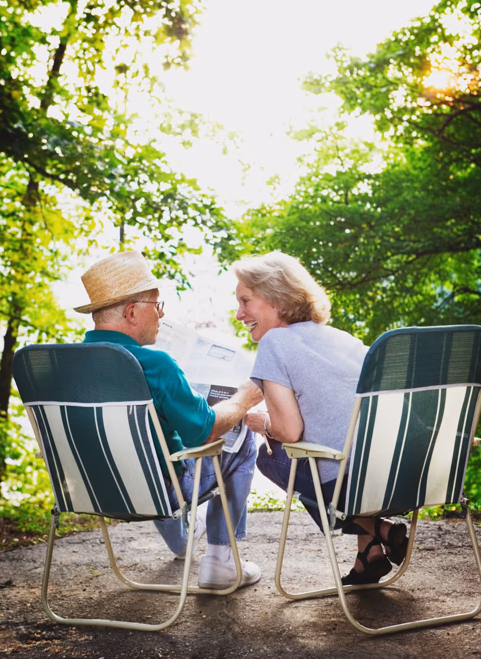Two seniors sit in striped outdoor chairs, talking and smiling among green trees.