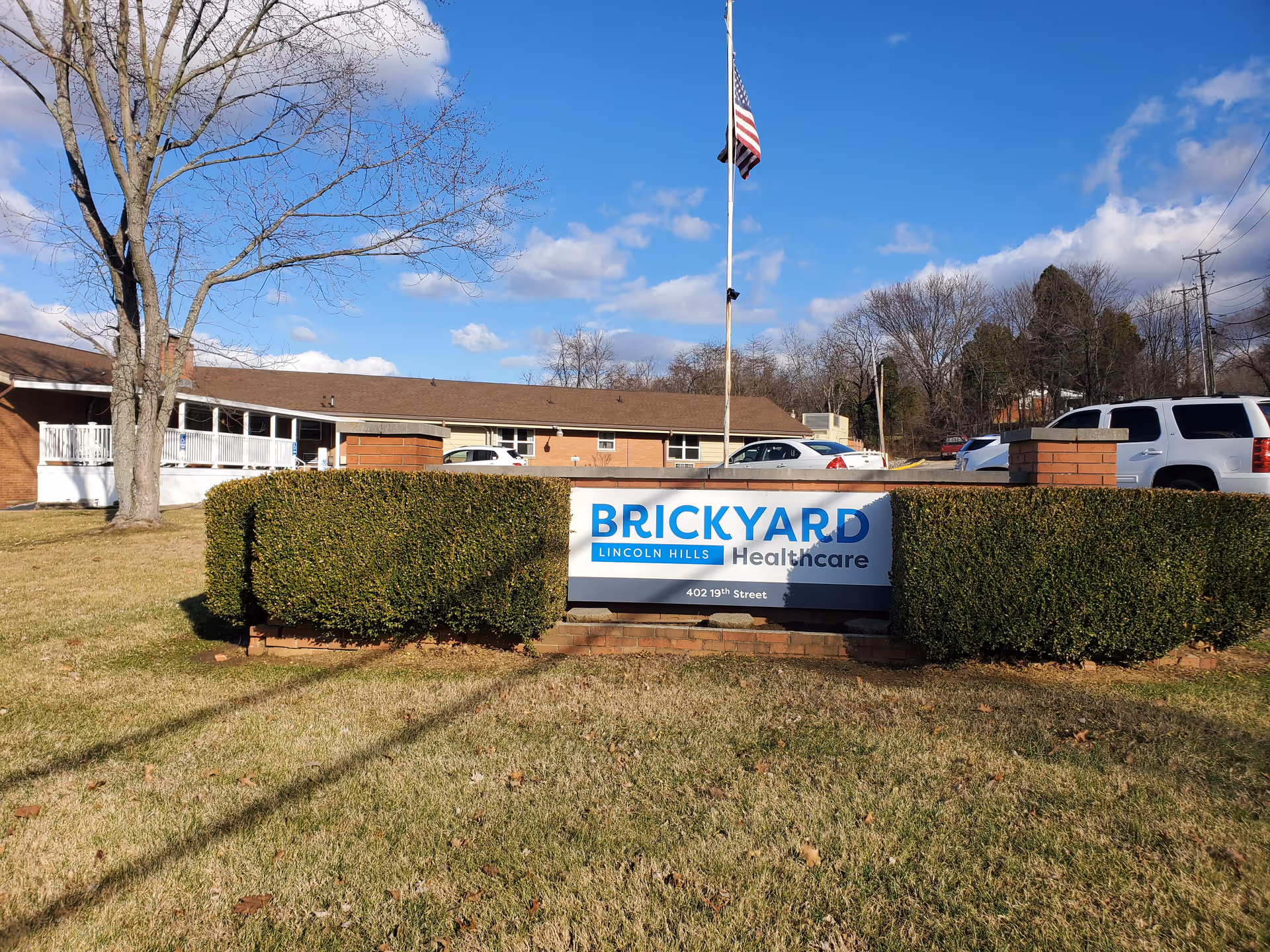Exterior view of a brick facility with Brickyard Healthcare sign, flagpole, hedges, and parked cars.