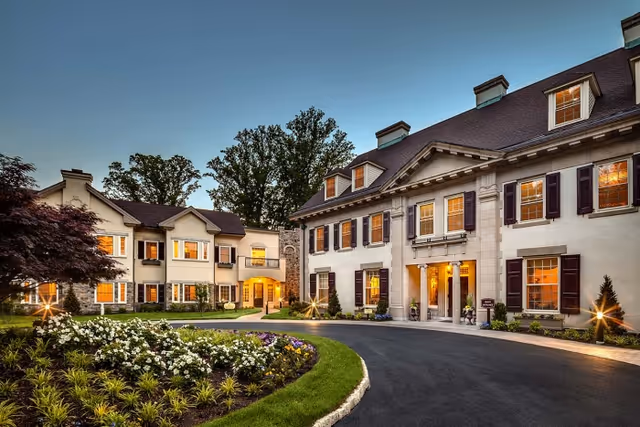 Entrance and circular driveway of a large, mansion-style senior living facility with lit windows, landscaped flower beds, and an evening sky.