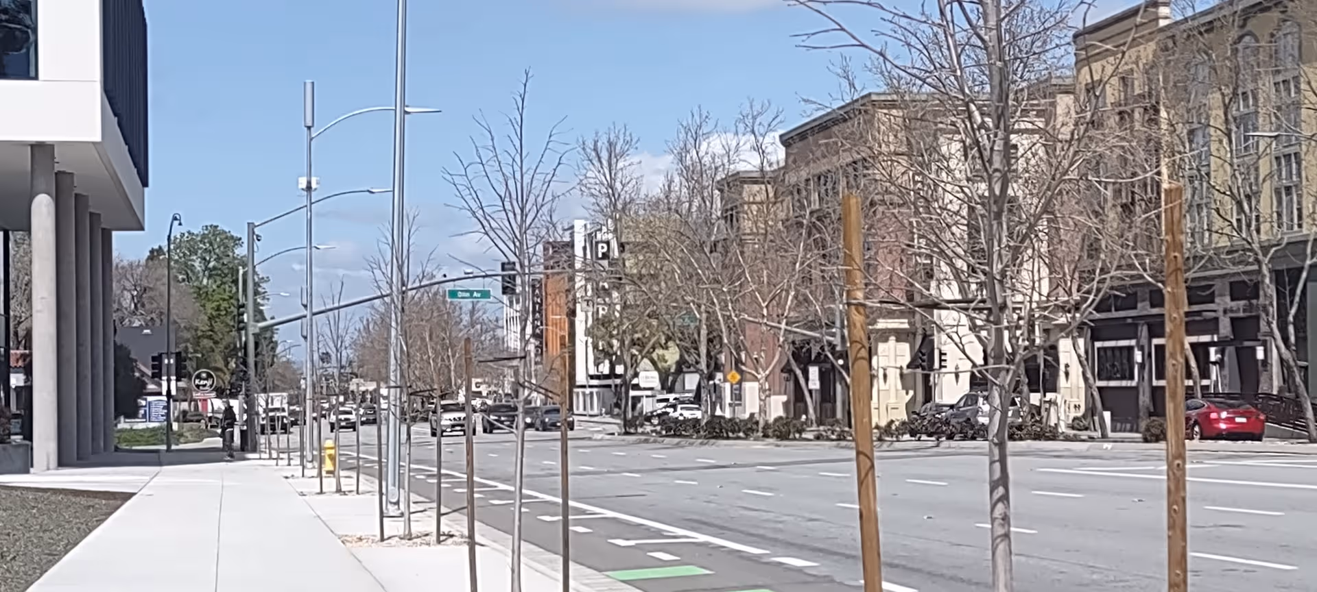 Urban street scene with modern building façade, bare trees, and empty sidewalk.