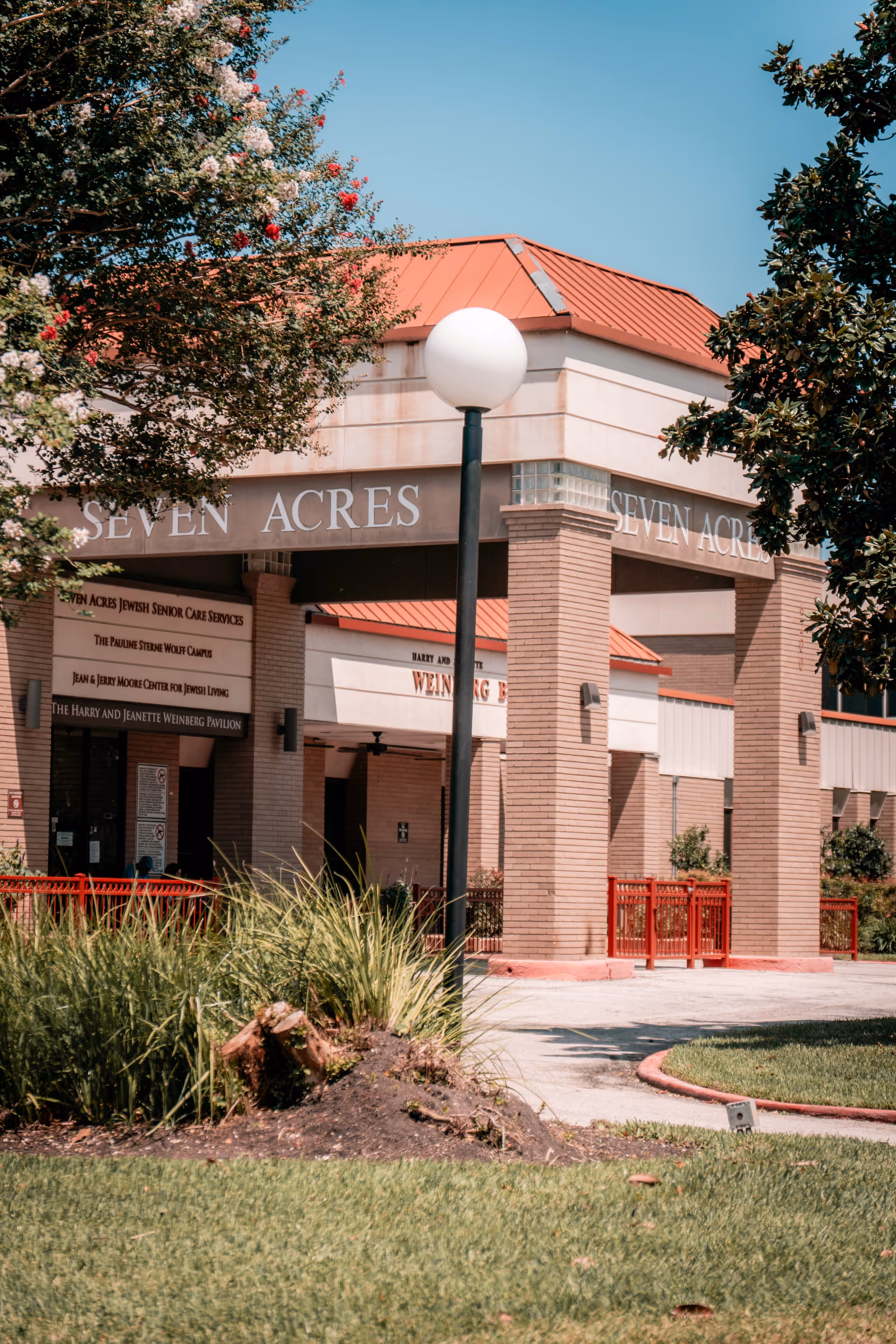 Exterior view of the entrance to Seven Acres Jewish Senior Care Services building with a red roof, brick pillars, a lamp post, and surrounding greenery including trees and grass.