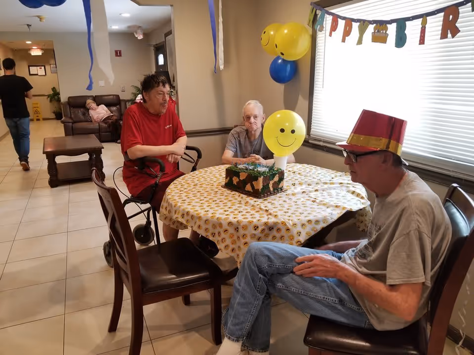 Three older adults sit around a decorated table with a cake and smiley balloons in a communal room celebrating a birthday.