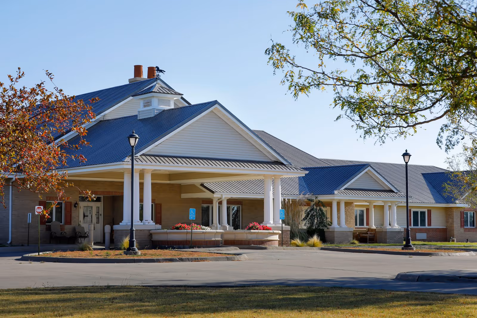 Exterior view of a single-story senior living facility building with a covered entrance supported by white columns, a blue metal roof, and surrounding trees and landscaping under a clear blue sky.