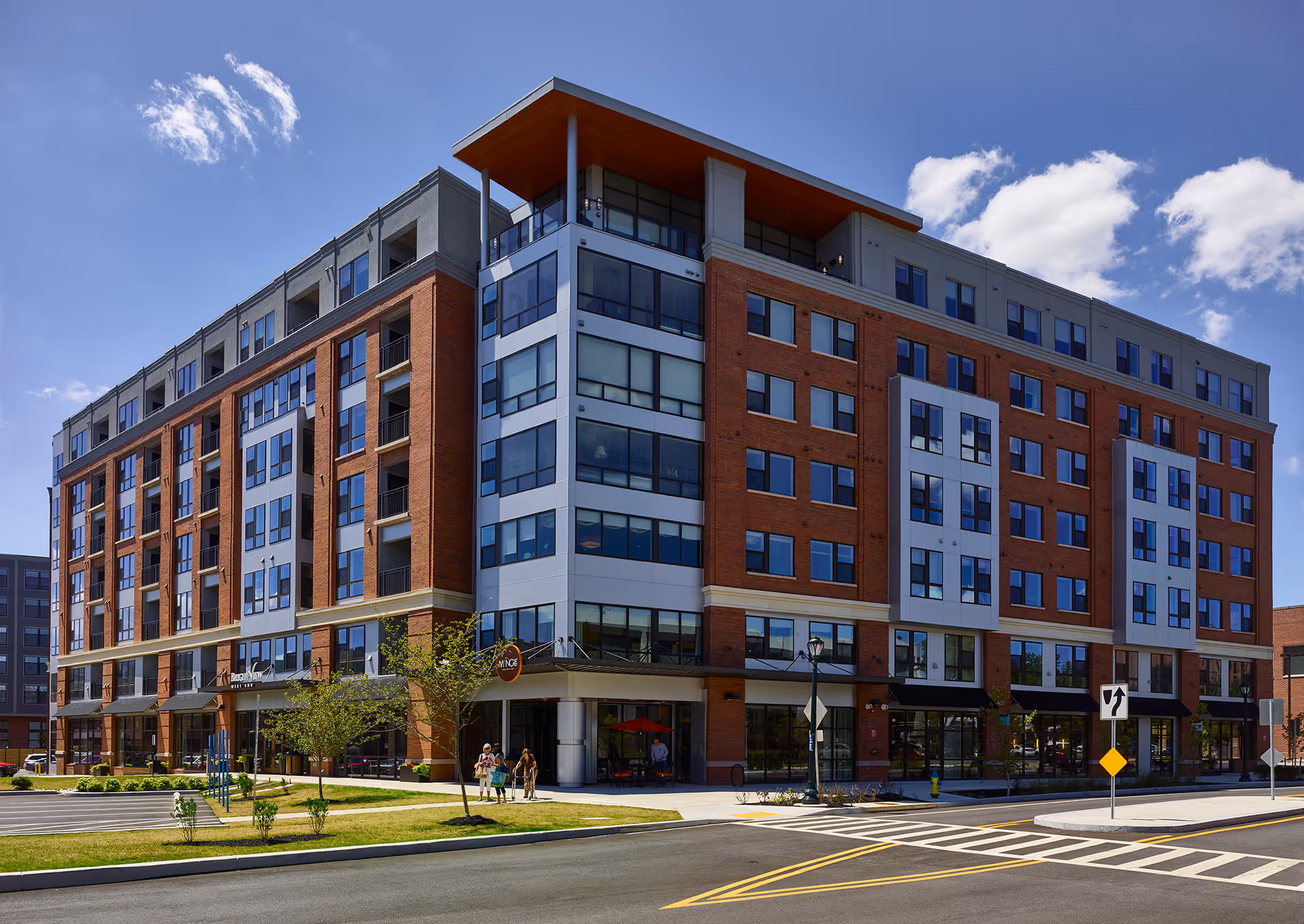 Exterior view of a modern multi-story senior living facility building with large windows, brick and gray paneling, a few small trees and grass areas, and a clear blue sky with some clouds. There are a few people walking near the entrance.