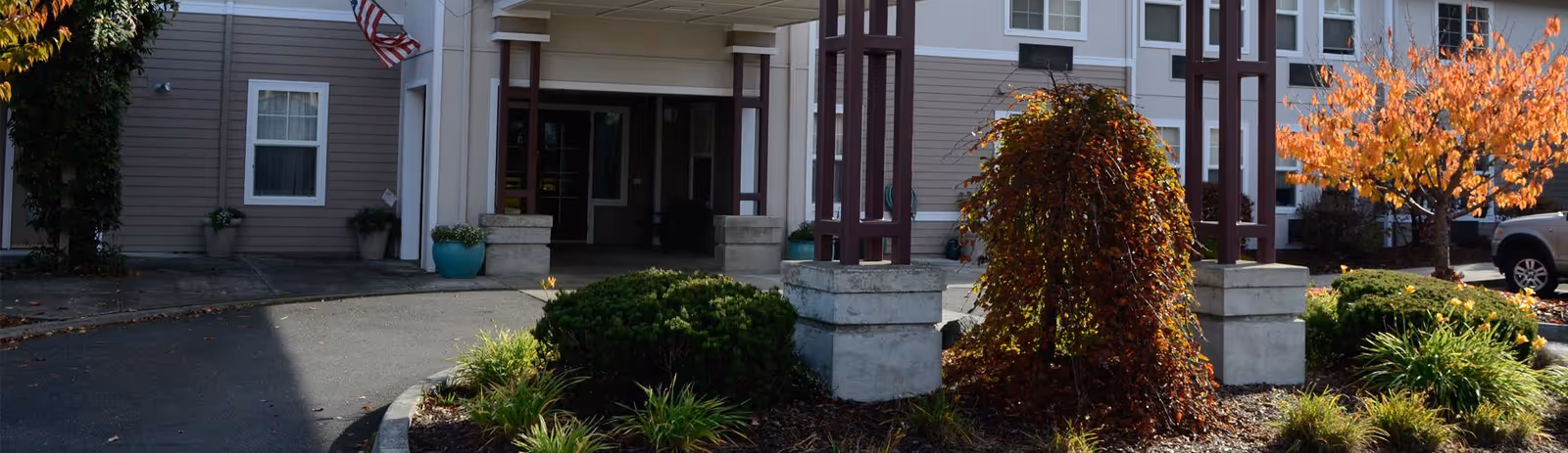 Covered main entrance of a senior living building with a porte-cochere, landscaped shrubs and fall-colored trees.
