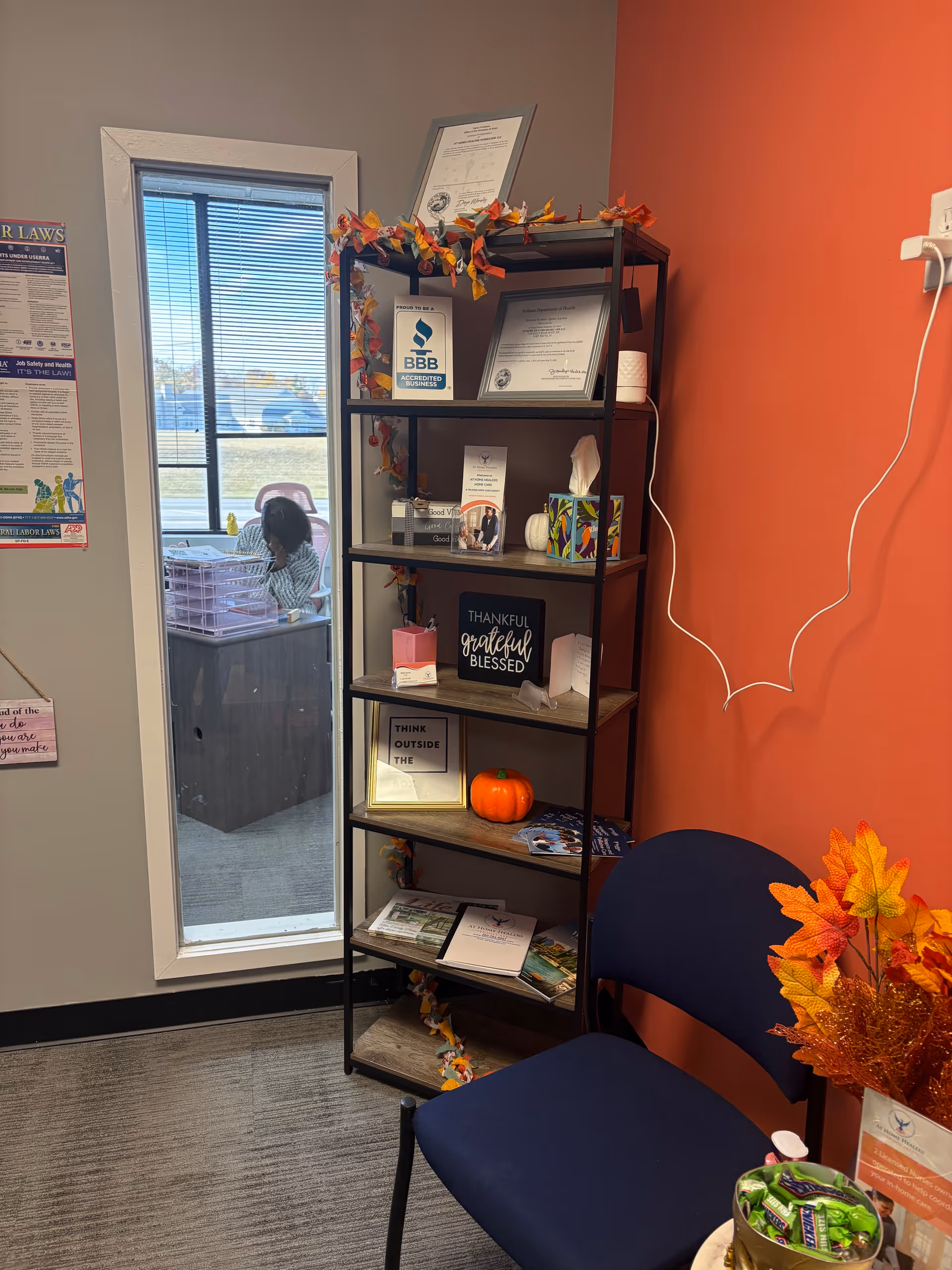 Tall metal shelf with decor, pumpkin, chair, and doorway to a room with a person.