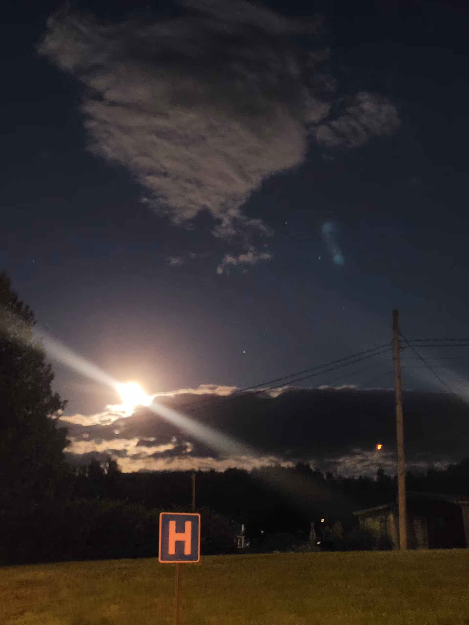 Night exterior of a hospital yard with an illuminated H sign and power lines.