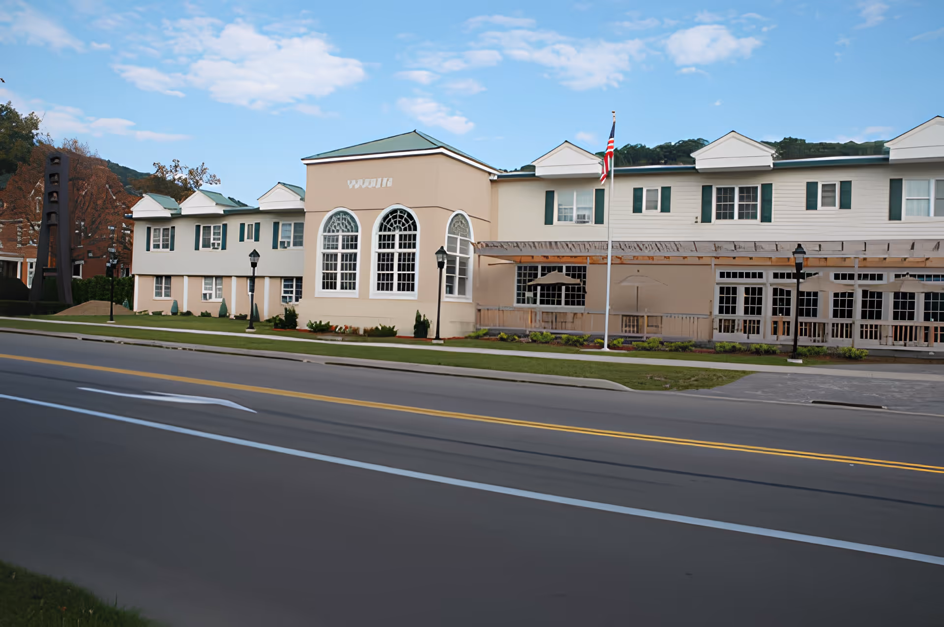 Exterior view of a senior living facility building with beige and white walls, green window shutters, and a green roof. The building features large arched windows and a covered patio area with umbrellas. An American flag is displayed on a flagpole in front of the building, and there is a road with marked lanes in the foreground. Trees and hills are visible in the background under a partly cloudy sky.