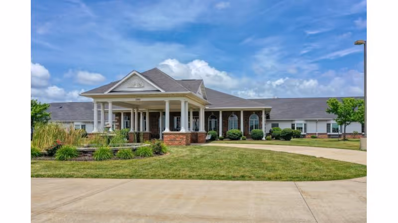Front exterior of a single-story brick senior living building with a covered portico, landscaped lawn, and blue sky.
