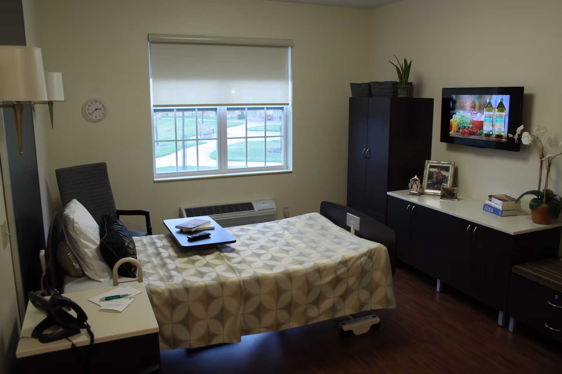 A small, neatly arranged bedroom in a skilled nursing facility featuring a single bed with a patterned bedspread, a tray table with a book and remote control, a cushioned chair, a wall-mounted TV, a cabinet with framed photos and books, a window with a roller blind, and a wall clock.