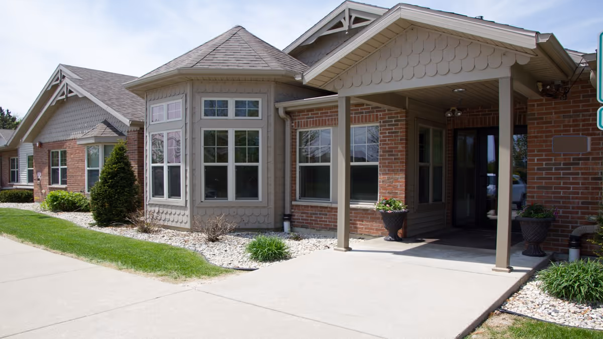 Exterior view of a single-story brick and siding building with a covered entrance supported by columns, landscaped with bushes and white rocks along the foundation, and a concrete walkway leading to the entrance.