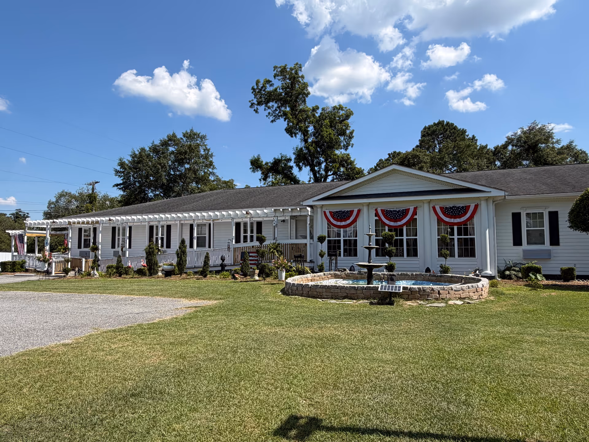 A single-story white senior living facility with a circular fountain, manicured lawn, and patriotic bunting.