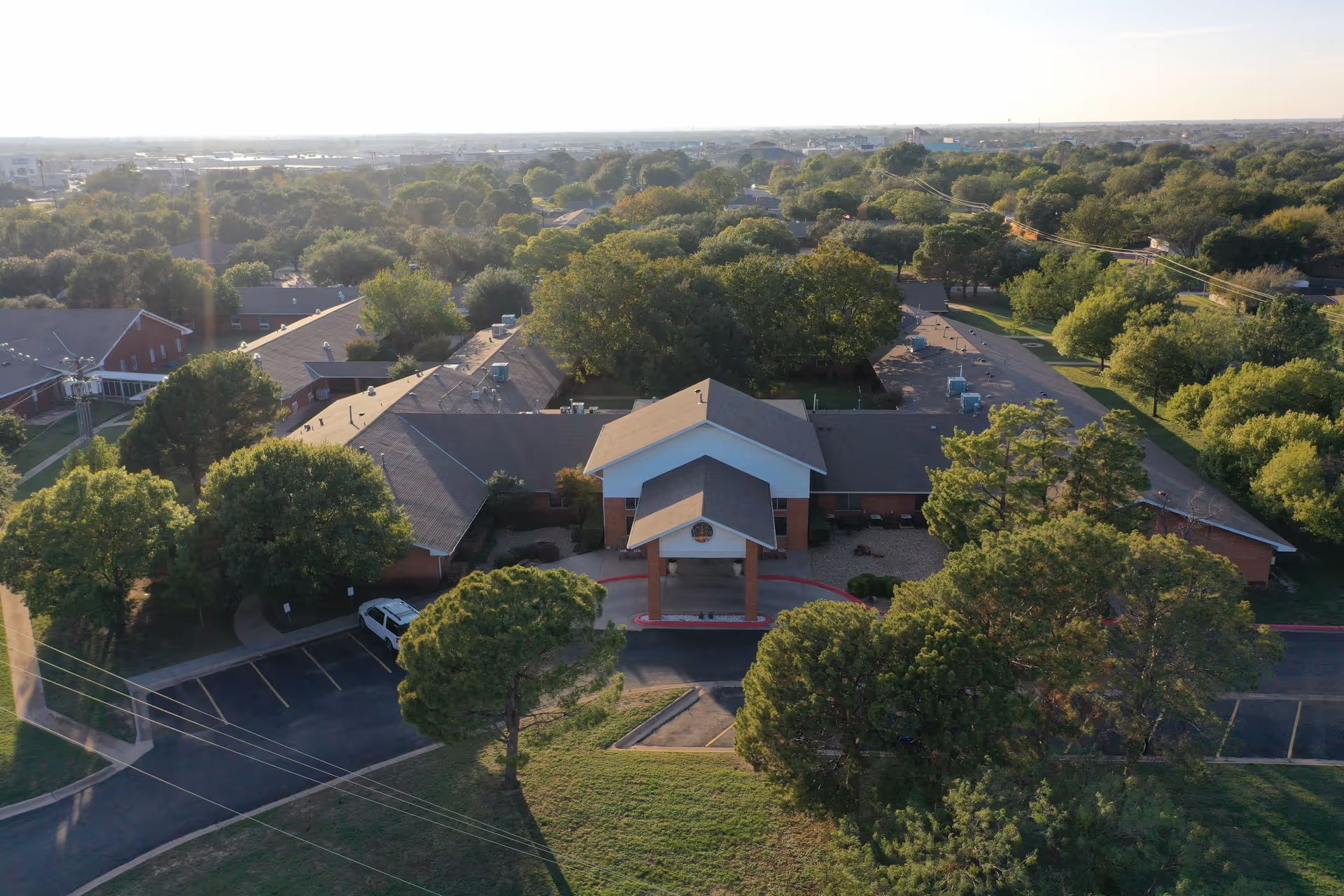 Aerial view of Wisteria Place Assisted Living facility showing a single-story building with a covered entrance, surrounded by trees and greenery under a clear sky.
