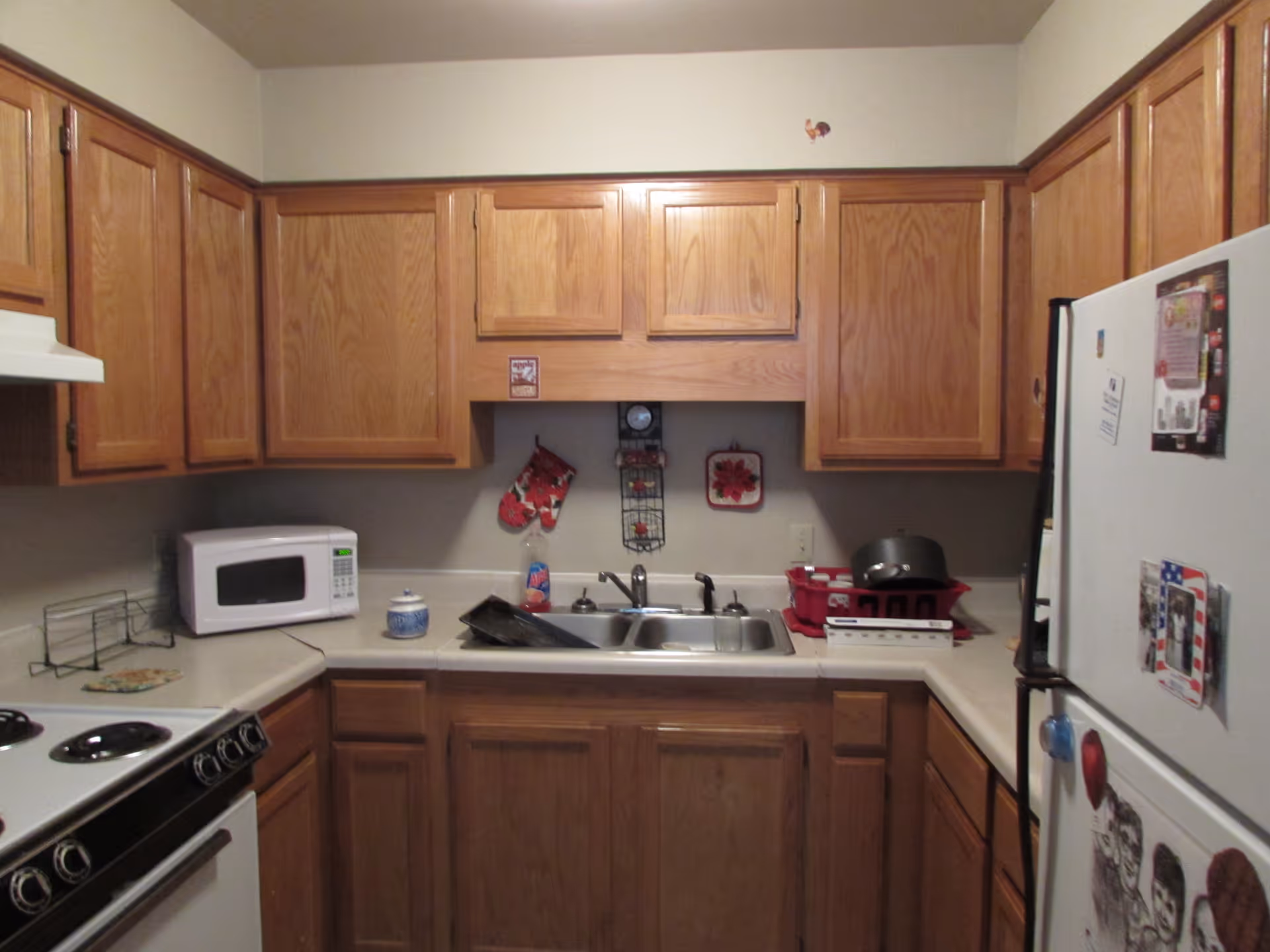 Compact kitchen with wooden cabinets, sink, microwave, stove, and refrigerator along L-shaped counters.