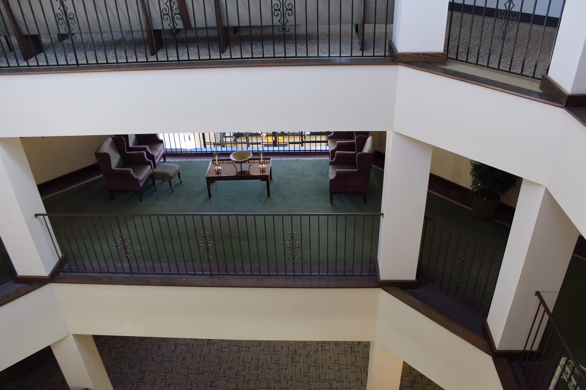View from an upper floor overlooking a seating area with two pairs of purple armchairs and a wooden coffee table on green carpet, surrounded by white walls and black metal railings.