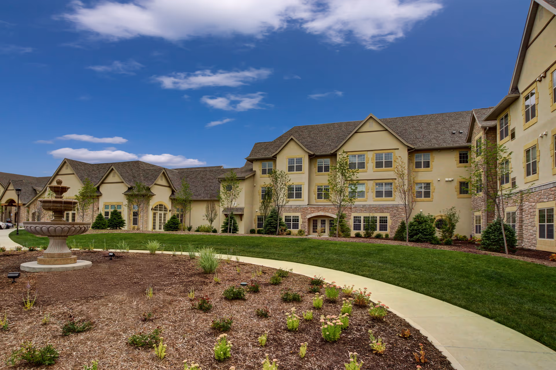 Exterior view of a senior living facility building with beige walls, multiple windows, and a sloped roof under a partly cloudy blue sky. In the foreground, there is a landscaped garden area with a stone fountain, young plants, and a curved concrete walkway surrounded by green grass.
