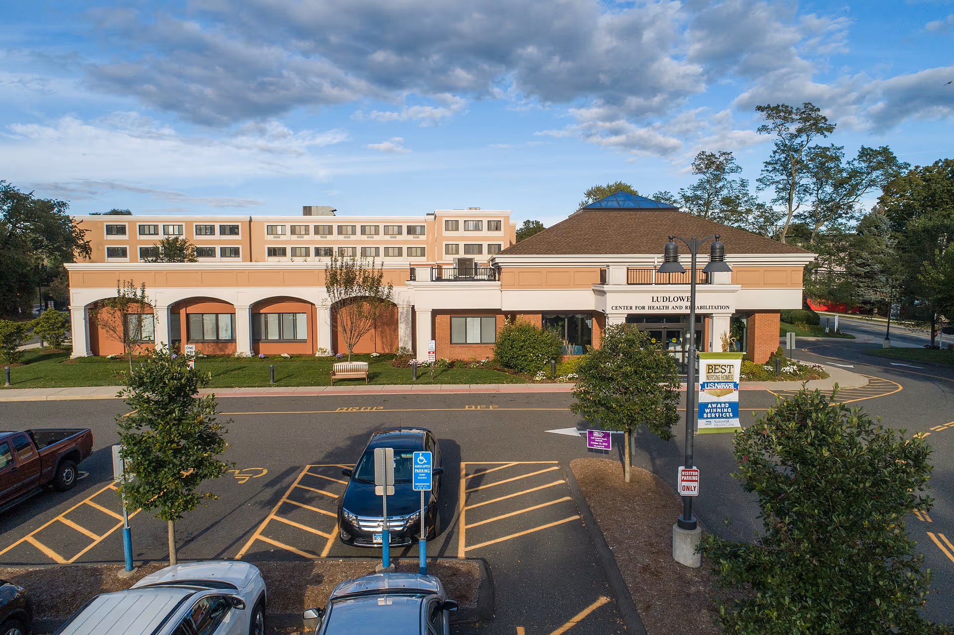 Exterior view of the Ludlowe Center for Health and Rehabilitation building with a parking lot in front, including handicap parking spaces and several cars parked. The building is a two-story structure with a beige and brick facade, surrounded by trees and greenery under a partly cloudy sky.