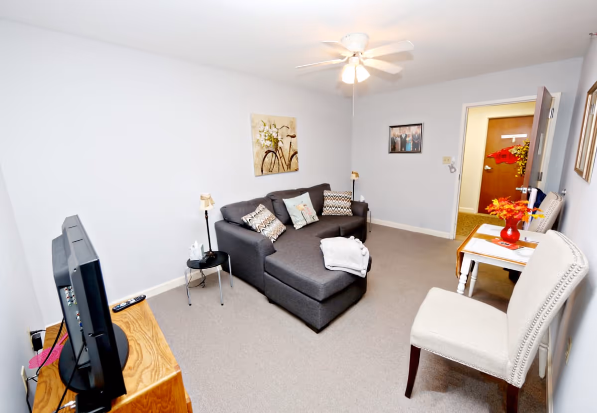 A small living room with a gray sectional sofa adorned with patterned pillows and a folded blanket. A small round side table with a lamp and tissue box sits next to the sofa. Across from the sofa is a TV on a wooden stand. There is a white upholstered chair and a small table with a red vase holding orange flowers. The walls are light gray with framed artwork and photographs. A ceiling fan with lights is mounted on the ceiling, and an open door leads to another room.