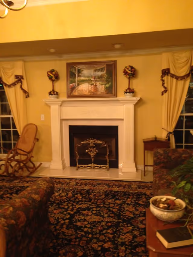 A cozy living room with a white fireplace centered on a yellow wall, flanked by two windows with yellow curtains tied back with maroon ribbons. Above the fireplace is a framed painting of a garden scene. On either side of the painting are decorative floral arrangements. In front of the fireplace is an ornate metal fireguard. To the left is a wooden rocking chair, and to the right is a small wooden side table. The floor is covered with a dark floral patterned carpet. A floral upholstered sofa is partially visible in the foreground along with a wooden table holding a bowl and a book.
