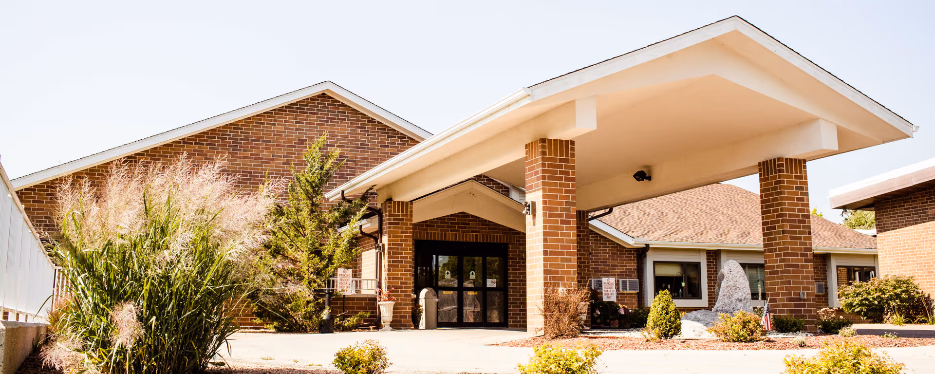 Front entrance of a brick senior living facility with a covered porte-cochere, glass doors, and landscaped shrubs.
