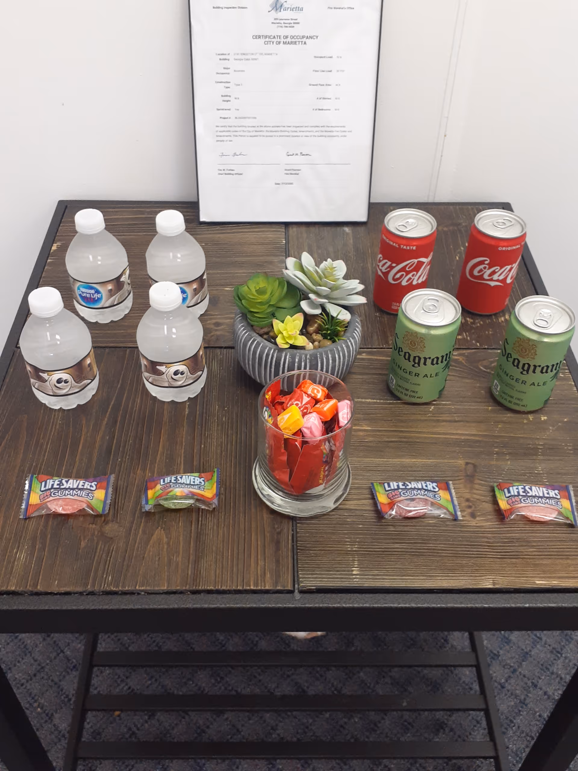 A wooden table displays water bottles, soda cans, candies, a succulent centerpiece, and a certificate indoors.